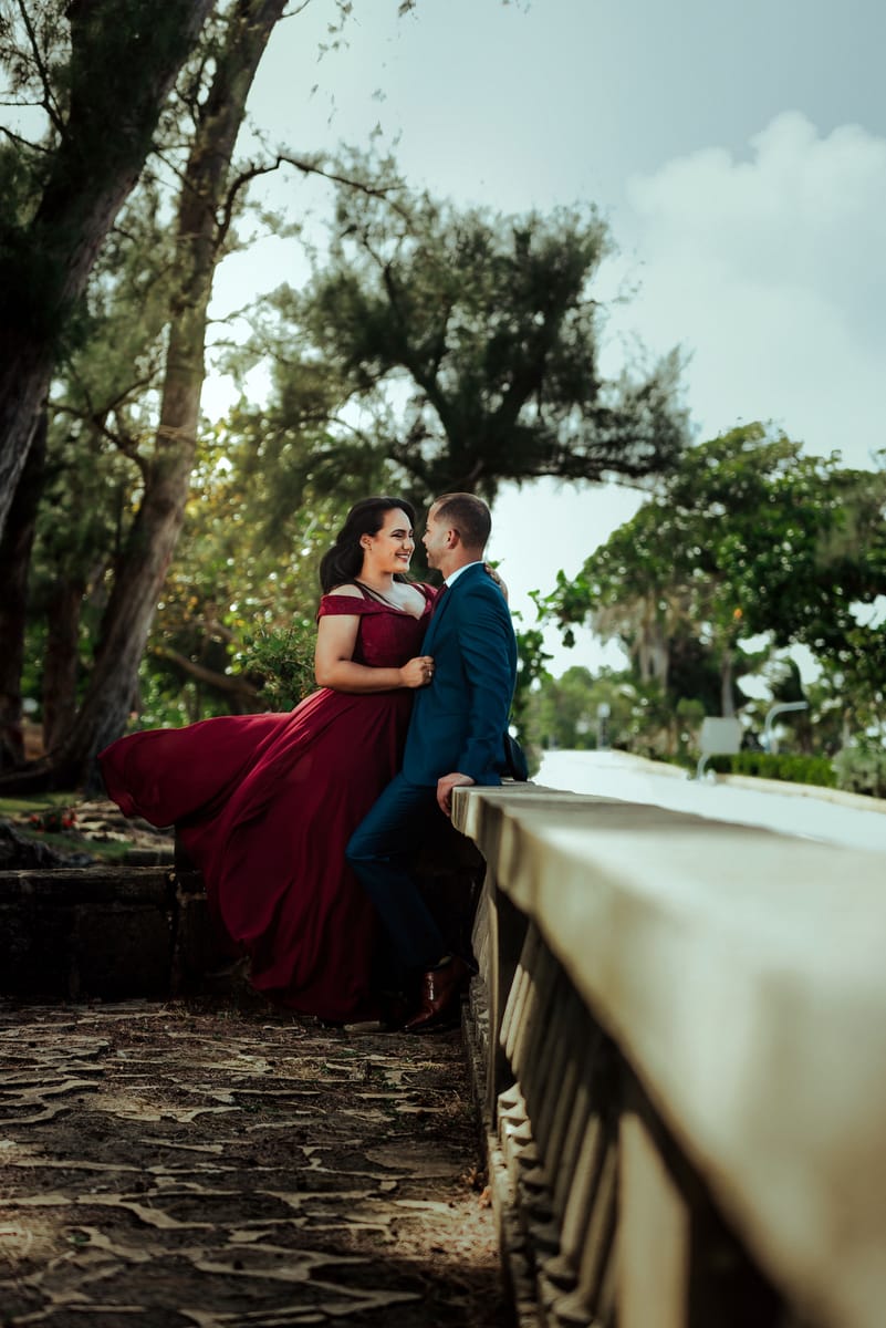 Pareja junto a una balaustrada de piedra, ella con vestido largo color vino que el viento mueve, entre árboles