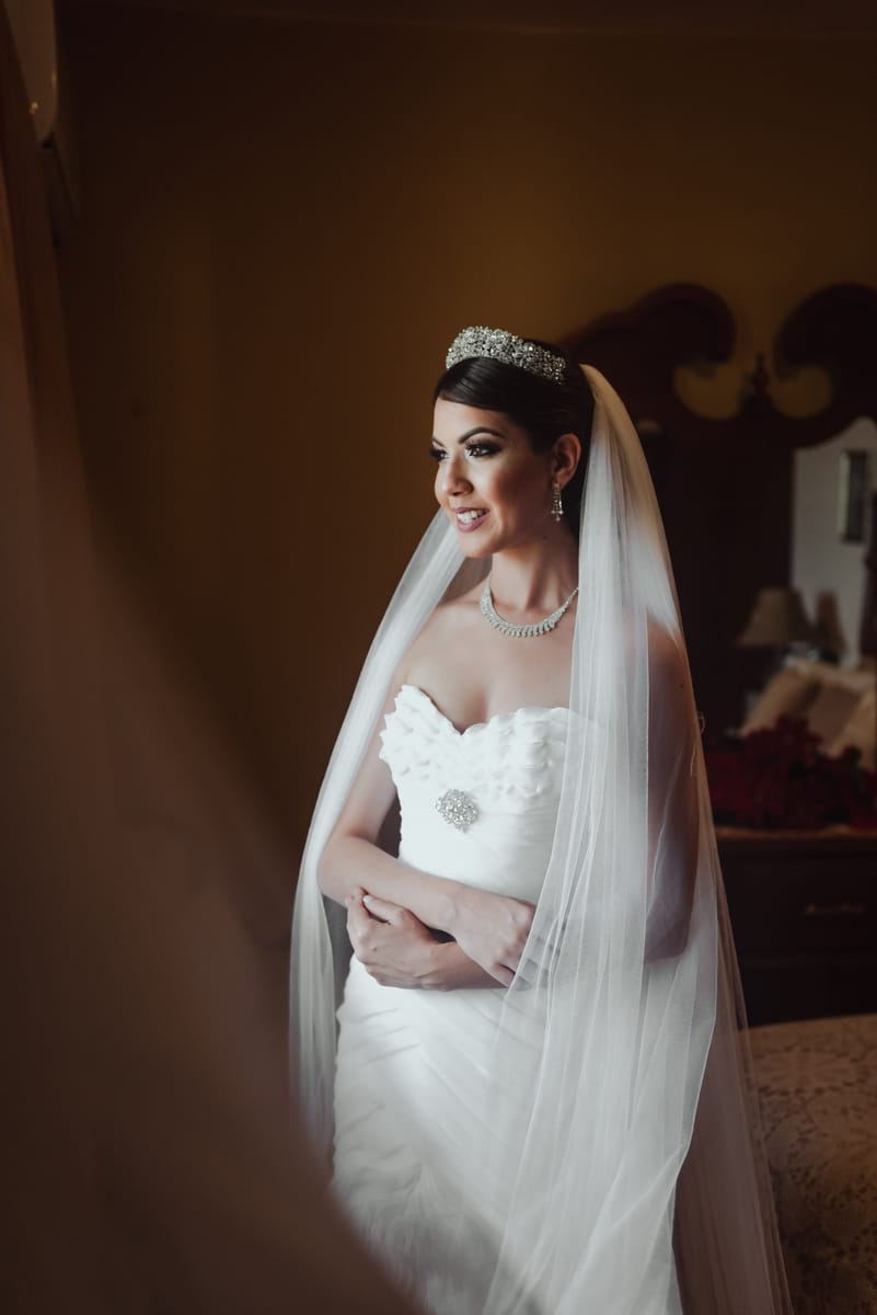 La novia con velo y tiara sonríe junto a una ventana, con luz suave