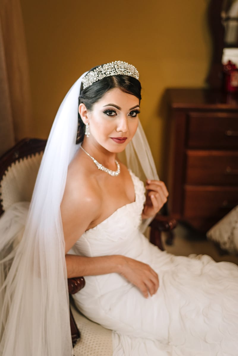 Retrato de la novia con velo y corona, sentada junto a una cómoda