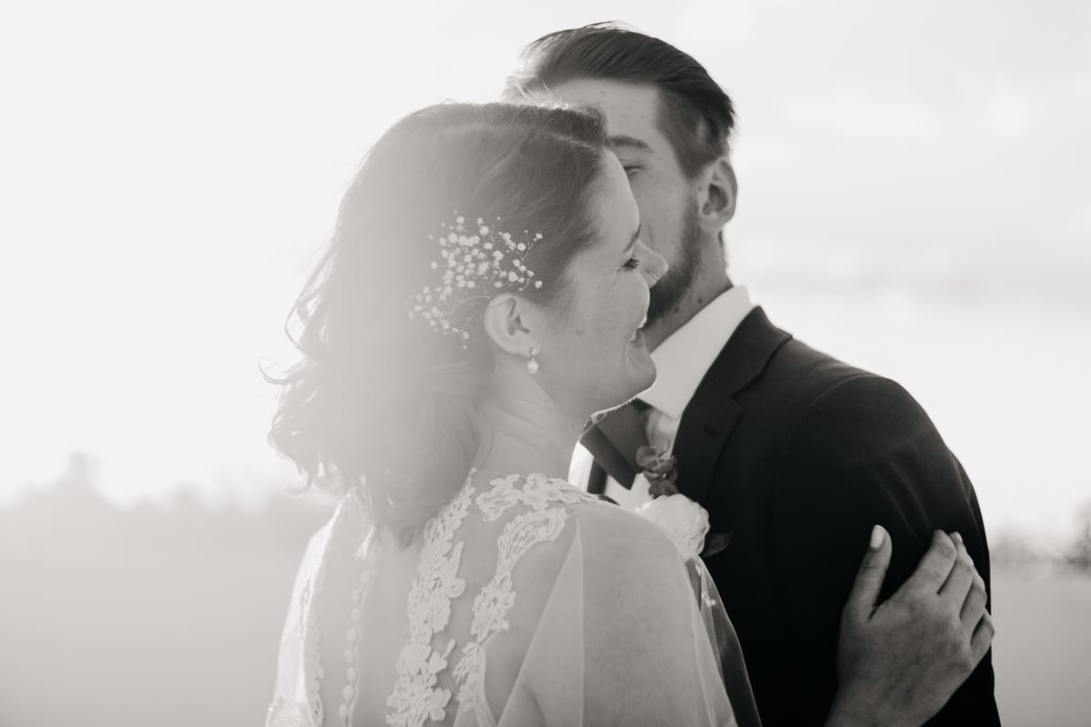 Retrato en blanco y negro de los novios a contraluz; él le besa la mejilla y ella ríe