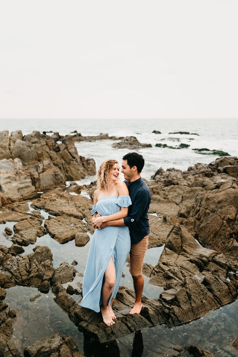 Pareja abrazada de pie sobre rocas con pozas de agua y el mar al fondo; ella viste un vestido azul claro