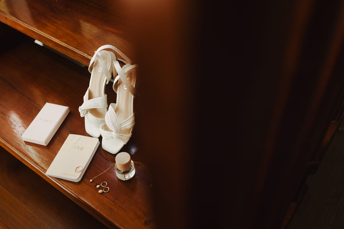 Zapatos blancos de la novia, un librito, joyas y perfume sobre un estante de madera
