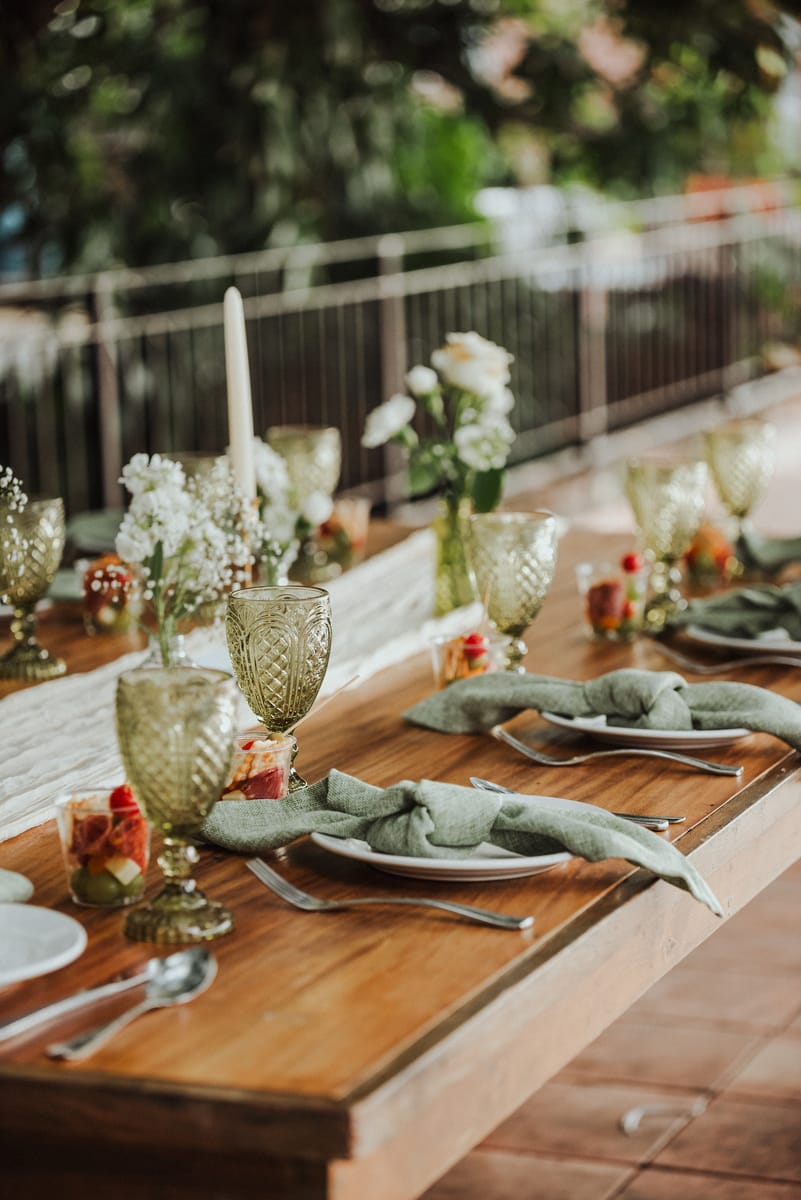 Mesa de boda de madera con copas verdes, servilletas de lino verde salvia, flores blancas y una vela