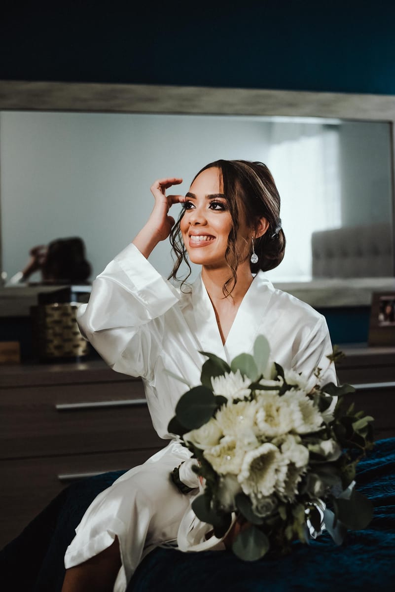 Novia en bata blanca sonríe y se acomoda el cabello con su ramo en las manos