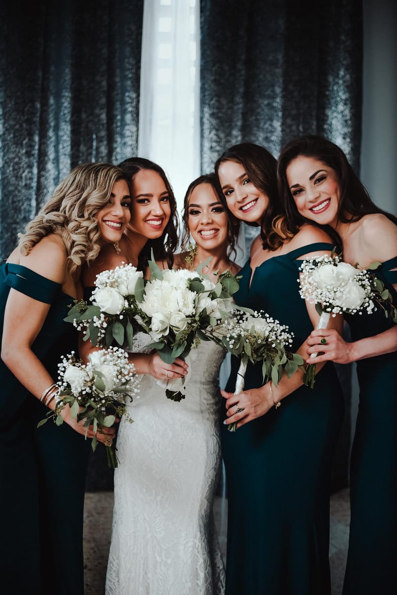 La novia abrazada por sus damas, de verde esmeralda, todas sonriendo con ramos blancos