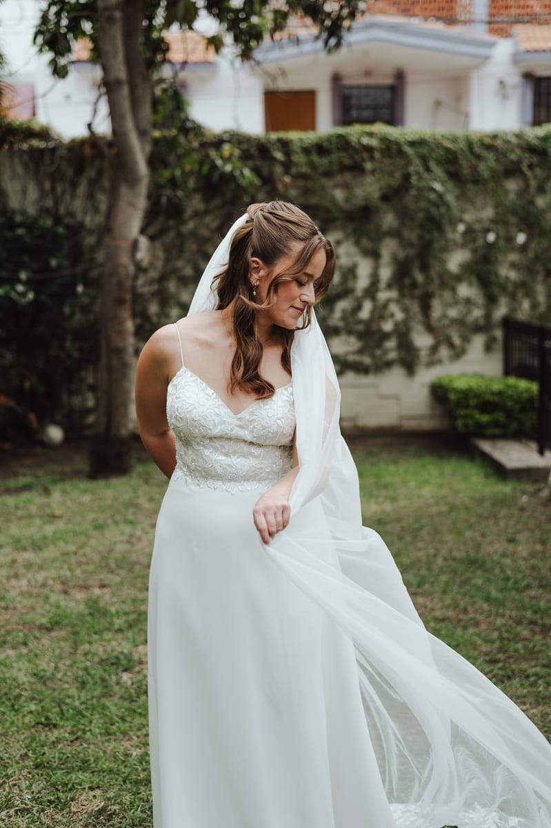 La novia en un jardín, con la mirada baja, sostiene con una mano el velo largo que el viento levanta
