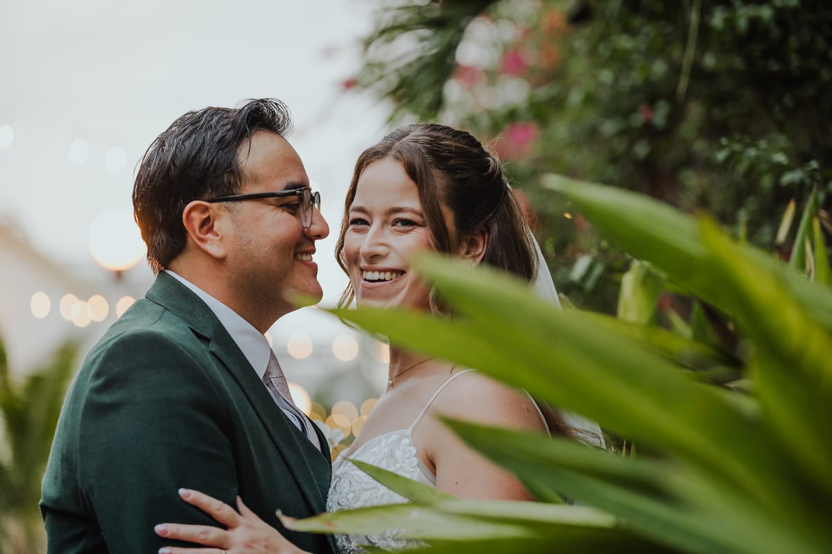 Los novios, él de traje verde y lentes, sonríen mirándose entre hojas verdes y luces desenfocadas