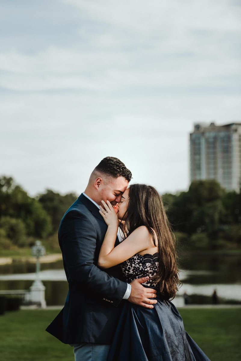 Pareja abrazada con las frentes juntas junto a un lago, con un edificio alto al fondo