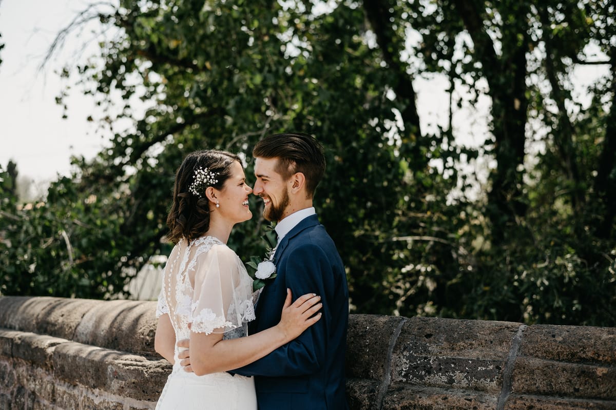 Los novios se miran sonriendo, frente a frente, junto a un muro de piedra y árboles