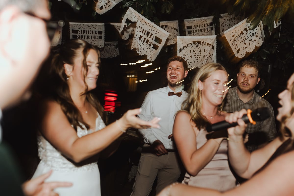 Invitados bailan y ríen en la fiesta bajo banderines de papel picado, con un micrófono en primer plano