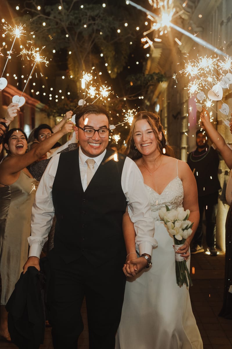 Los novios caminan de la mano y sonríen entre invitados que alzan luces de bengala