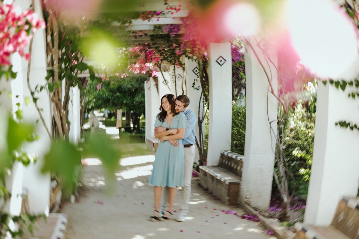 Pareja abrazada y sonriendo bajo un paseo de columnas blancas cubierto de flores rosas, con pétalos en el suelo