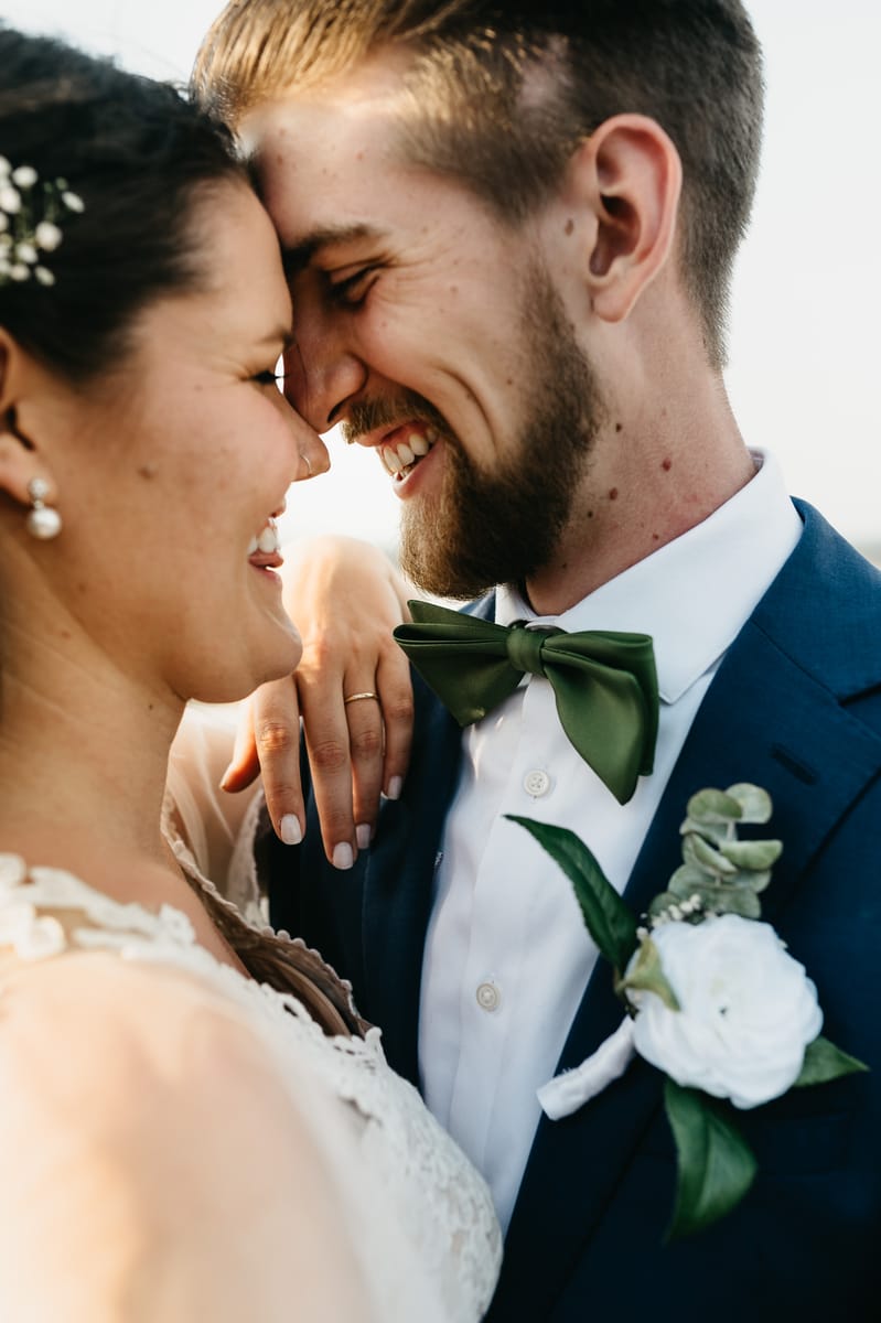Primer plano de los novios riendo con las frentes juntas; él lleva moño verde y una flor blanca en la solapa