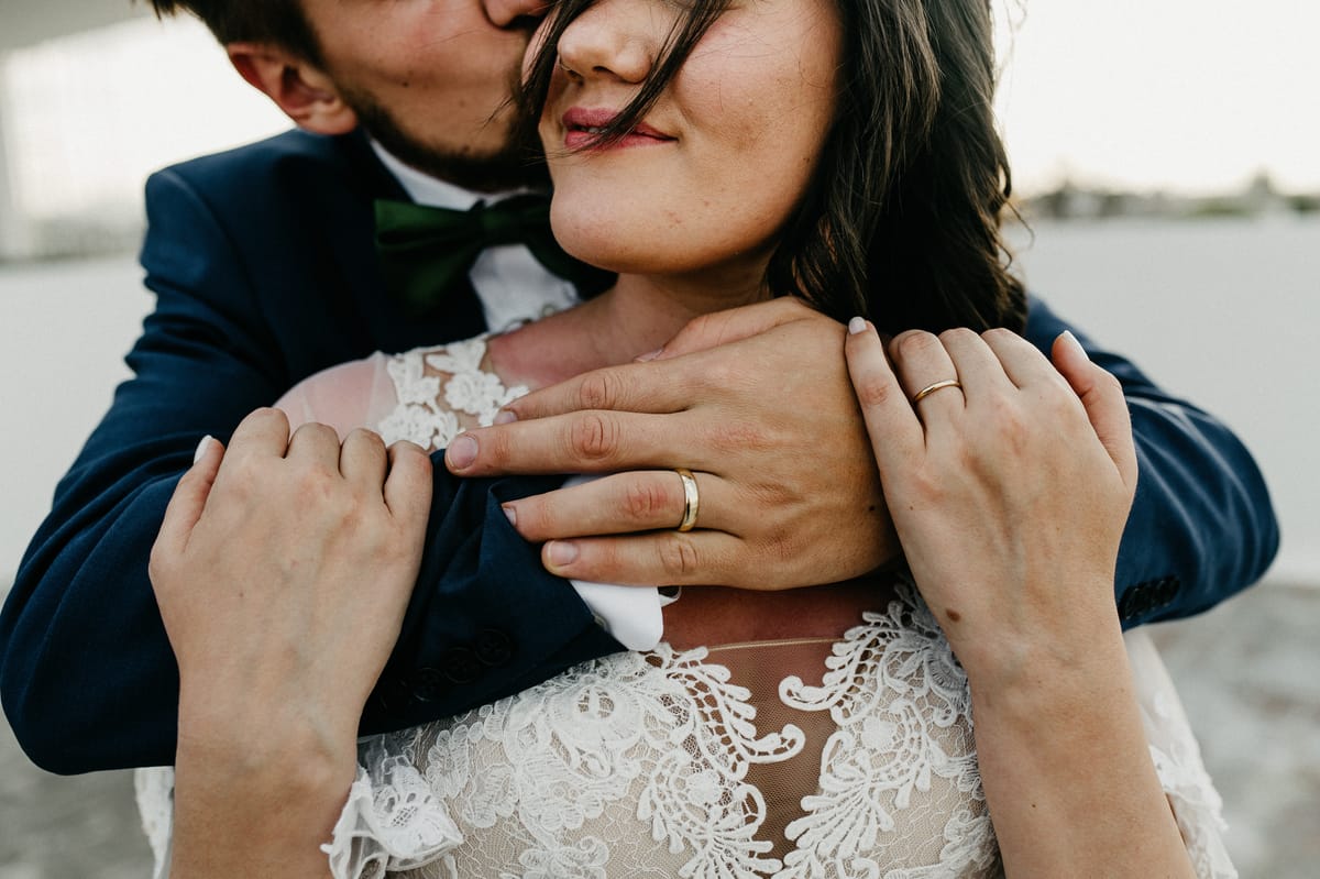 Detalle de las manos de los novios con sus alianzas sobre el encaje del vestido, mientras él la besa en la mejilla