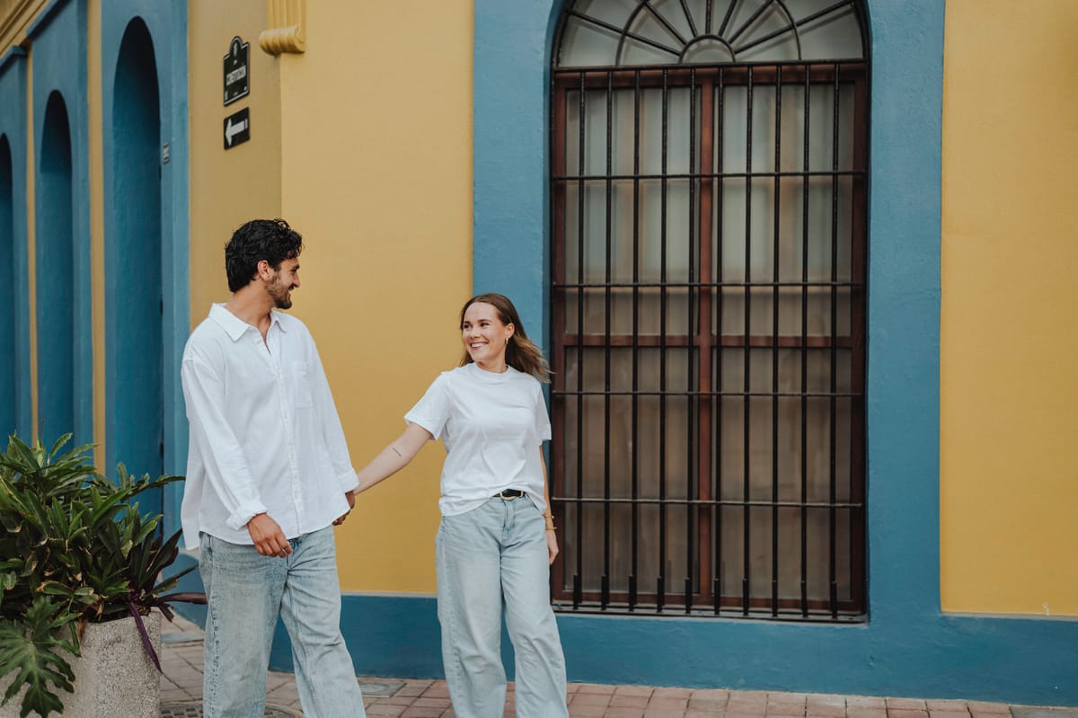 Pareja de la mano caminando frente a una pared amarilla y azul con una gran ventana enrejada