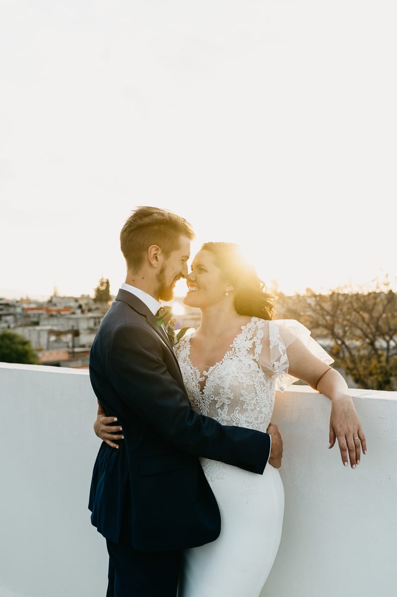 Los novios se abrazan a contraluz en una azotea al atardecer, con la ciudad al fondo