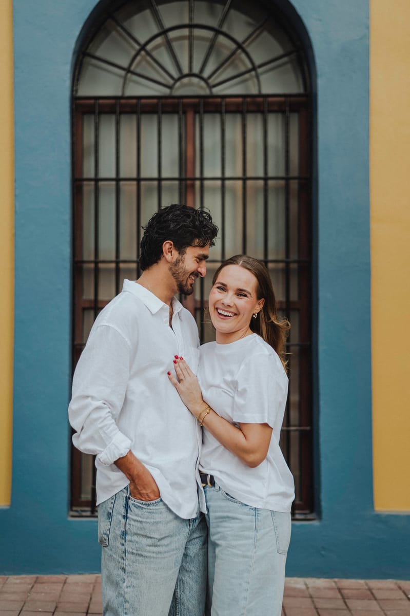 Pareja de blanco riendo abrazada frente a una ventana de reja con arco y un muro azul