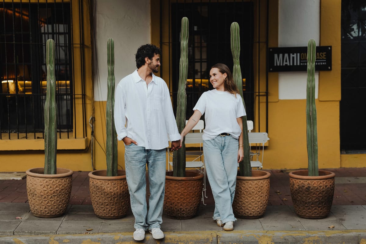 Pareja de la mano y mirándose entre grandes cactus en macetas de barro, frente a una fachada amarilla