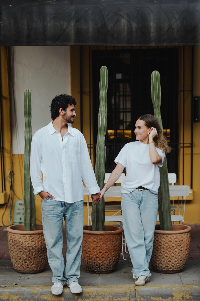 Pareja de la mano entre cactus altos en macetas de barro; ella se acomoda el cabello y lo mira