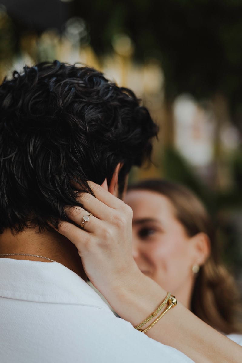 Detalle de la mano de ella, con un anillo, tocando el cabello de él; su sonrisa se ve desenfocada al fondo