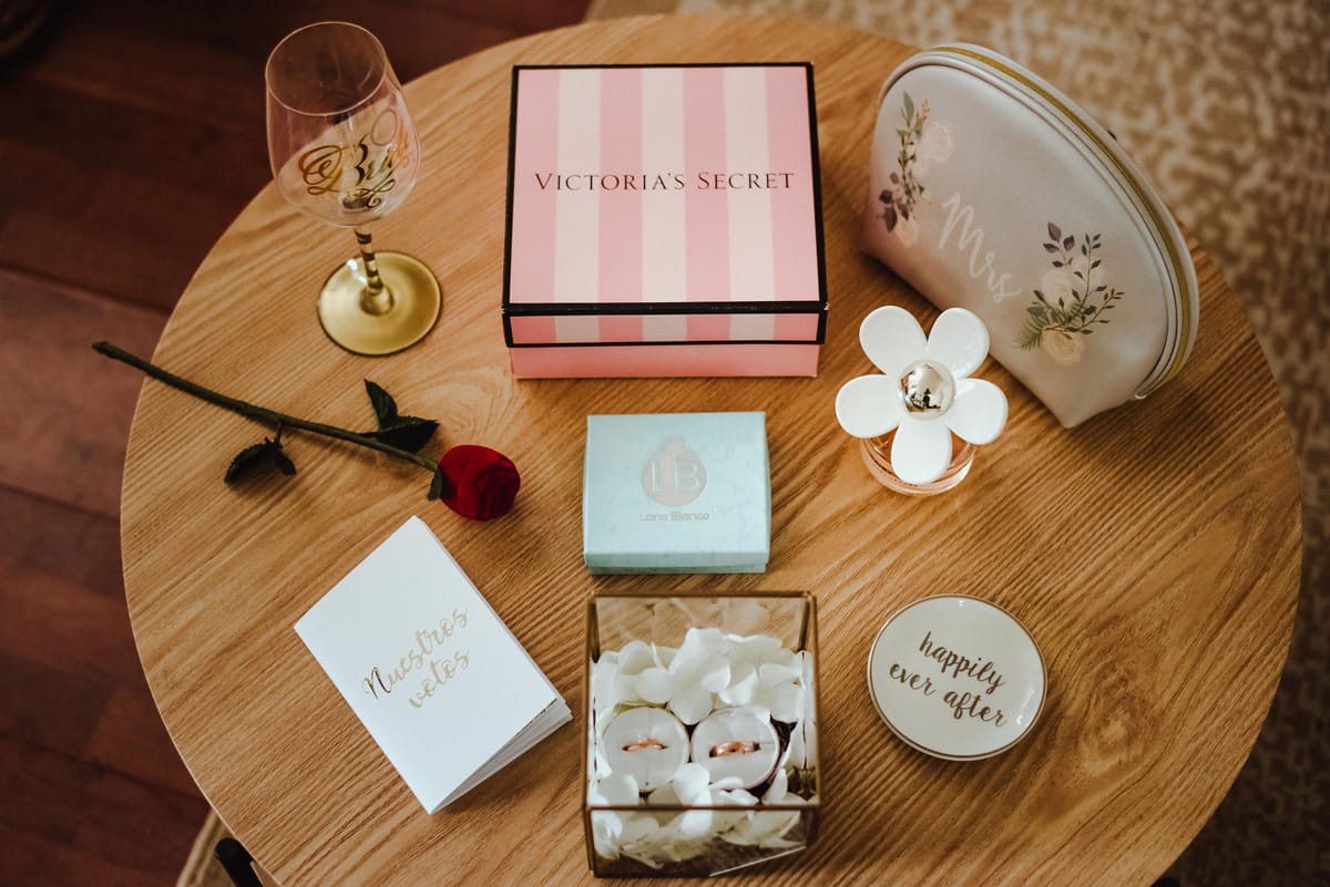 Mesa redonda con los detalles de la novia: una copa, una caja de regalo, una rosa, un librito de votos y los anillos en una caja de cristal