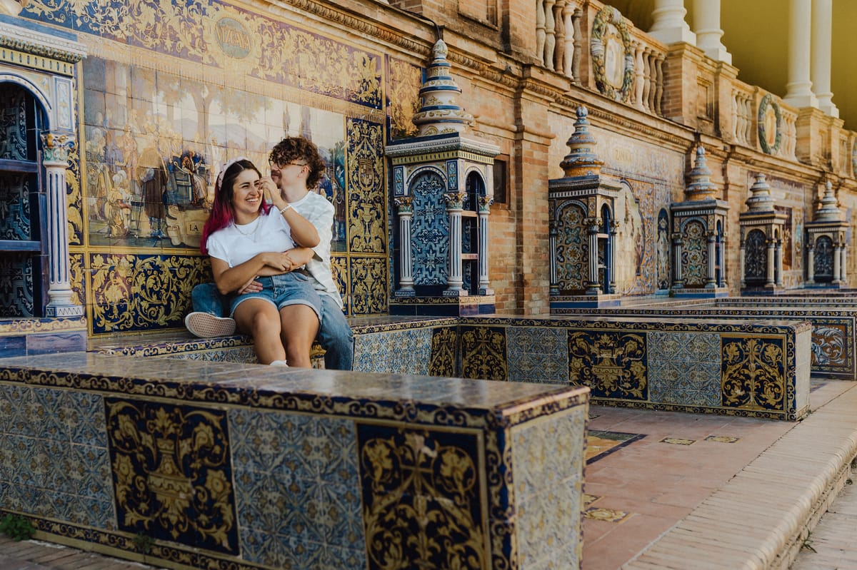 Pareja sentada abrazada en una banca decorada con azulejos, con un muro de azulejos pintados al fondo