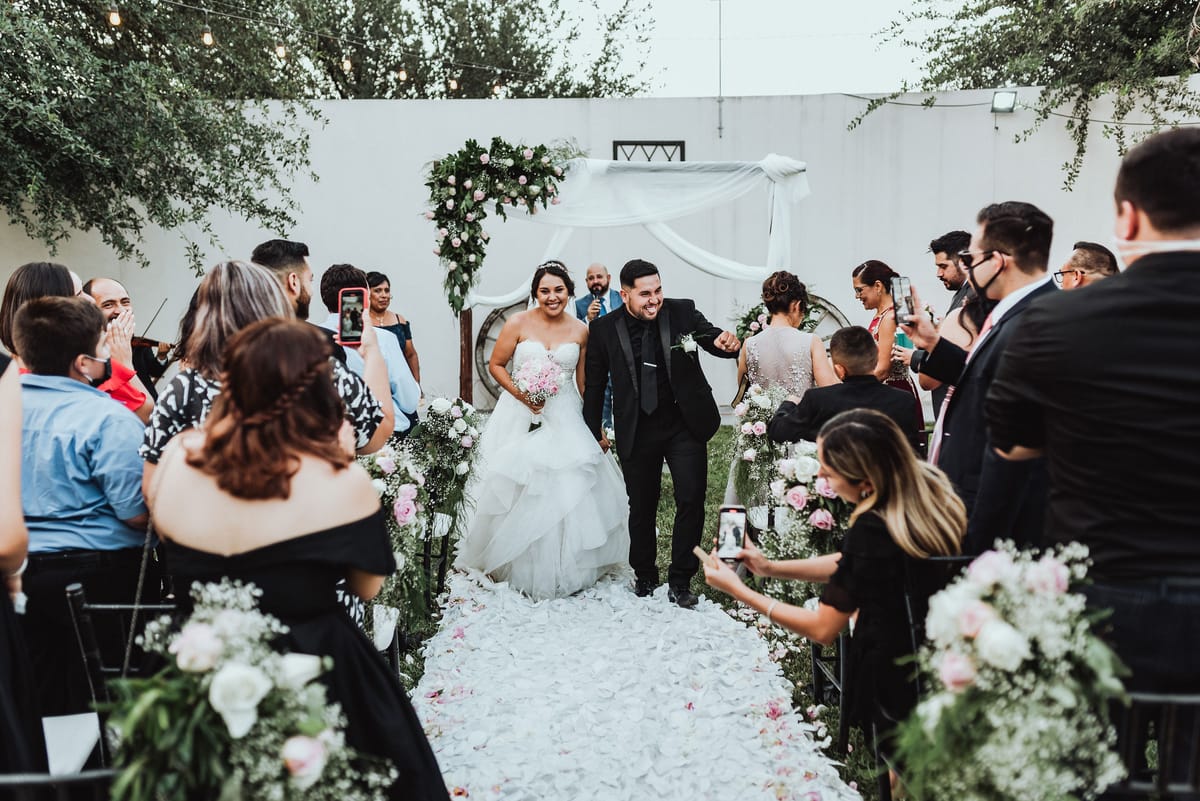 Los novios avanzan sonriendo por un pasillo de pétalos blancos, entre invitados que los fotografían