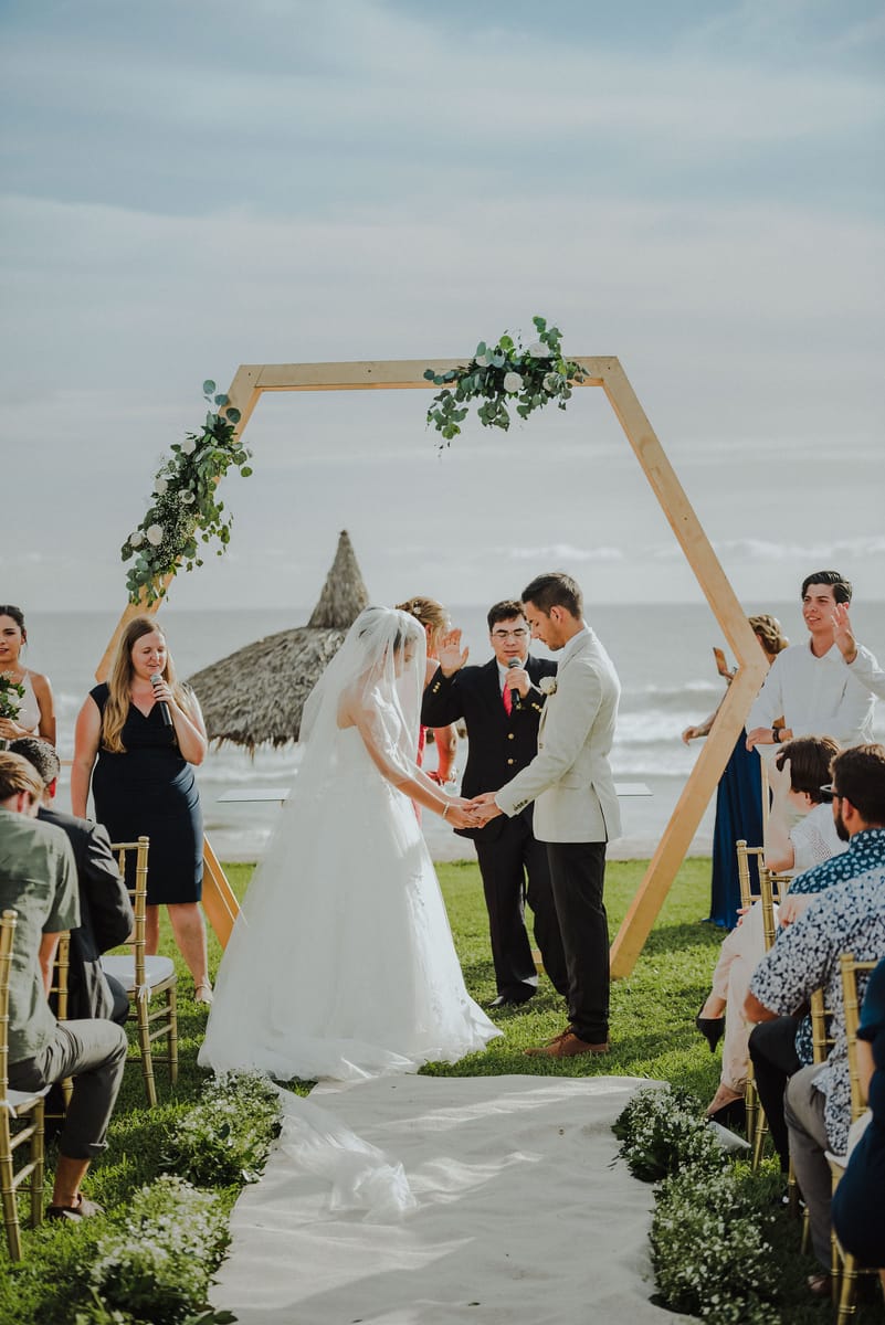 Los novios se toman de las manos bajo un arco hexagonal de madera frente al mar, con invitados sentados a los lados