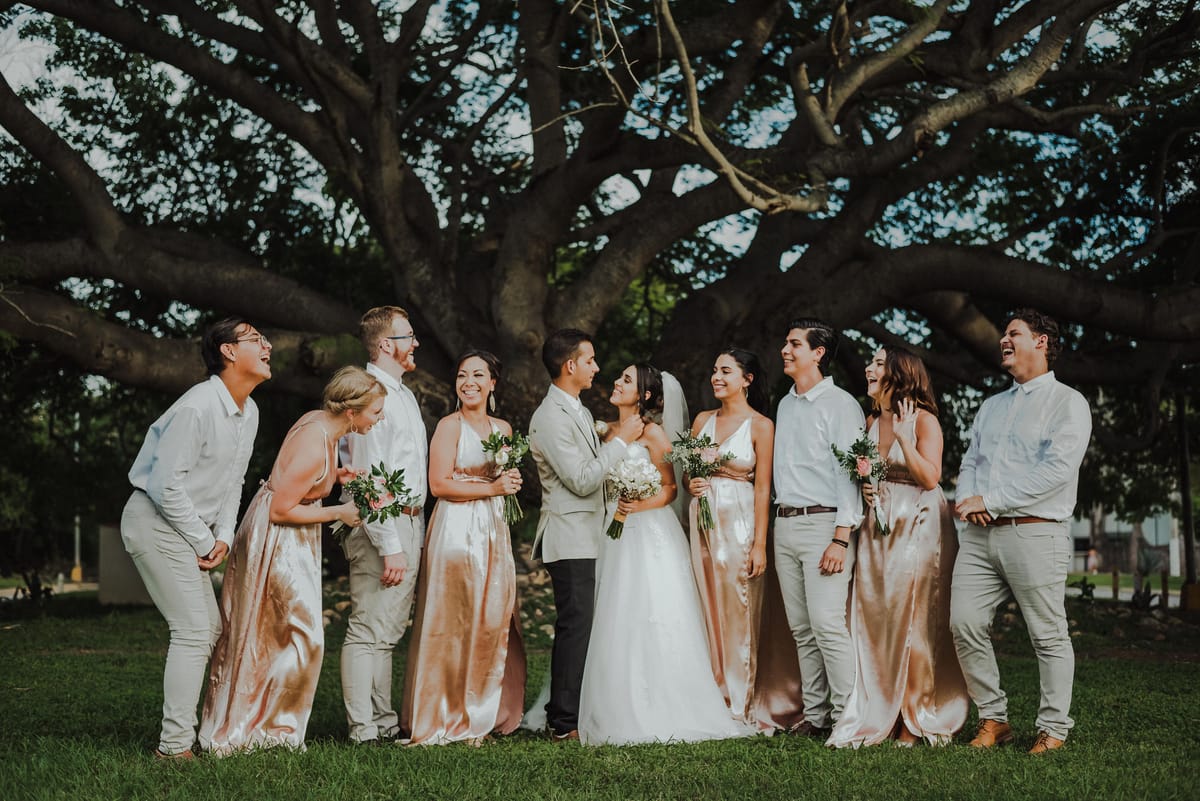 Los novios y su cortejo ríen juntos, de pie bajo un árbol enorme
