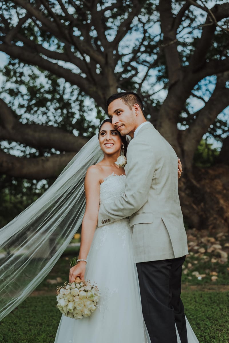 Los novios abrazados bajo un árbol; ella lleva el velo largo y un ramo de rosas blancas