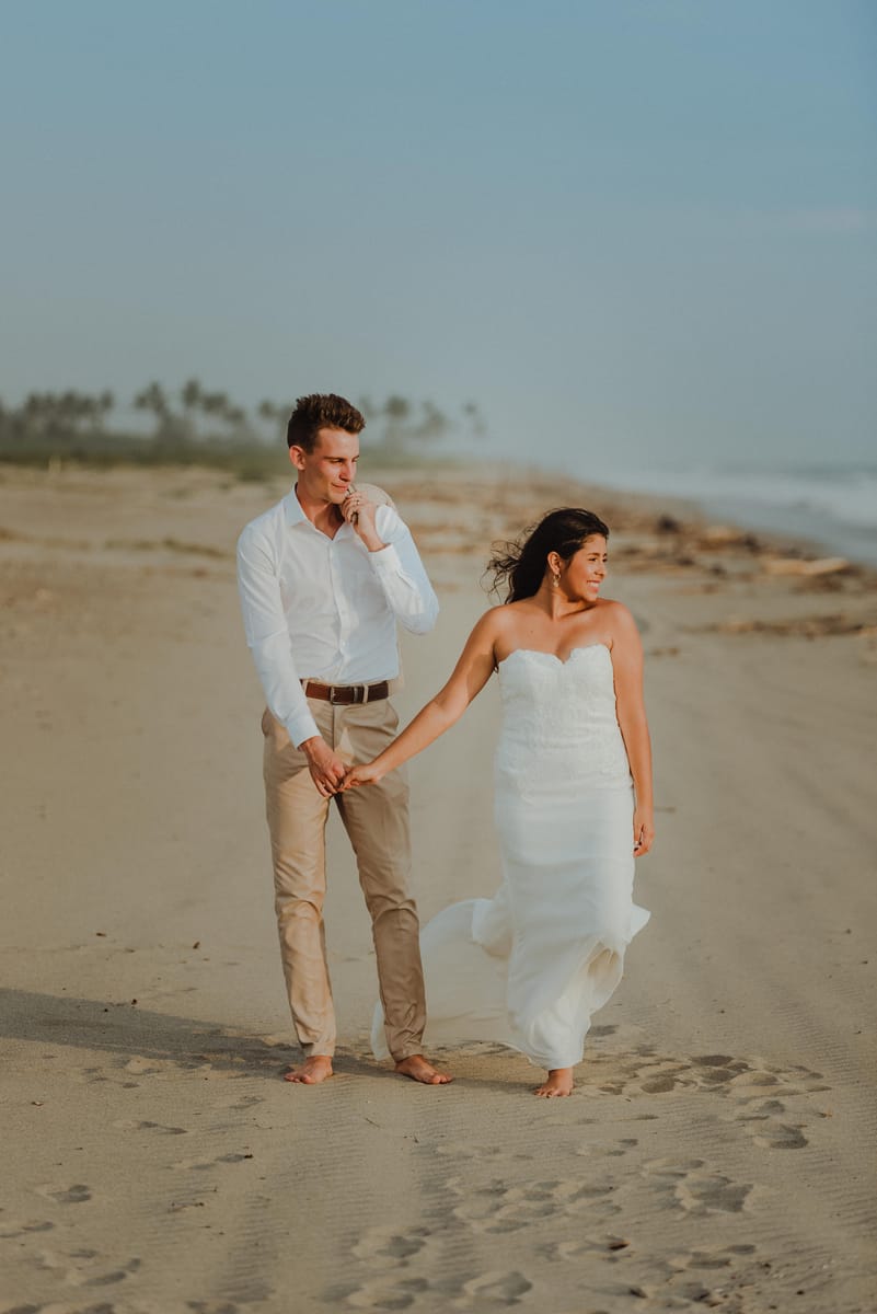 Los novios caminan de la mano descalzos por una playa; ella mira a lo lejos y él se lleva la mano a la boca