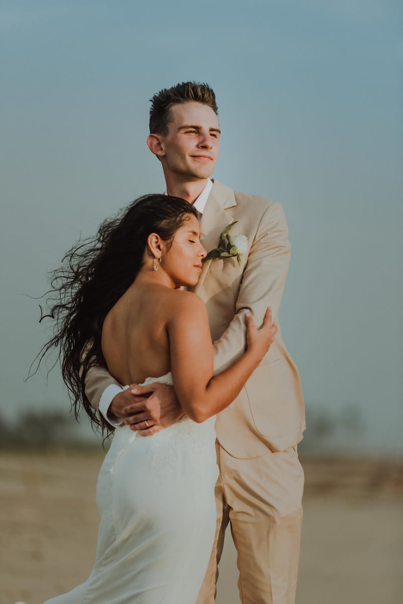 Los novios abrazados en la playa; ella, con el cabello al viento y los ojos cerrados, apoya la cabeza en su pecho