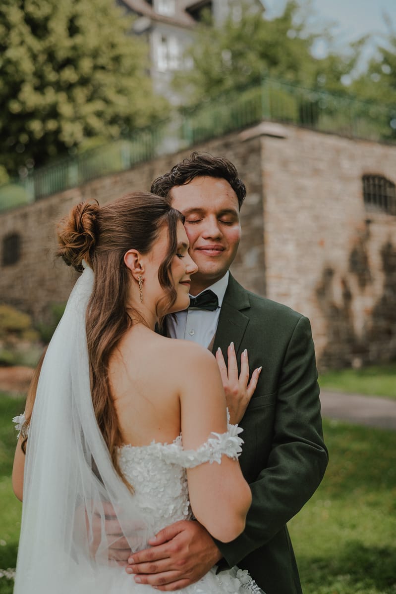 Retrato cercano de los novios abrazados con los ojos cerrados; él viste traje verde oscuro y moño