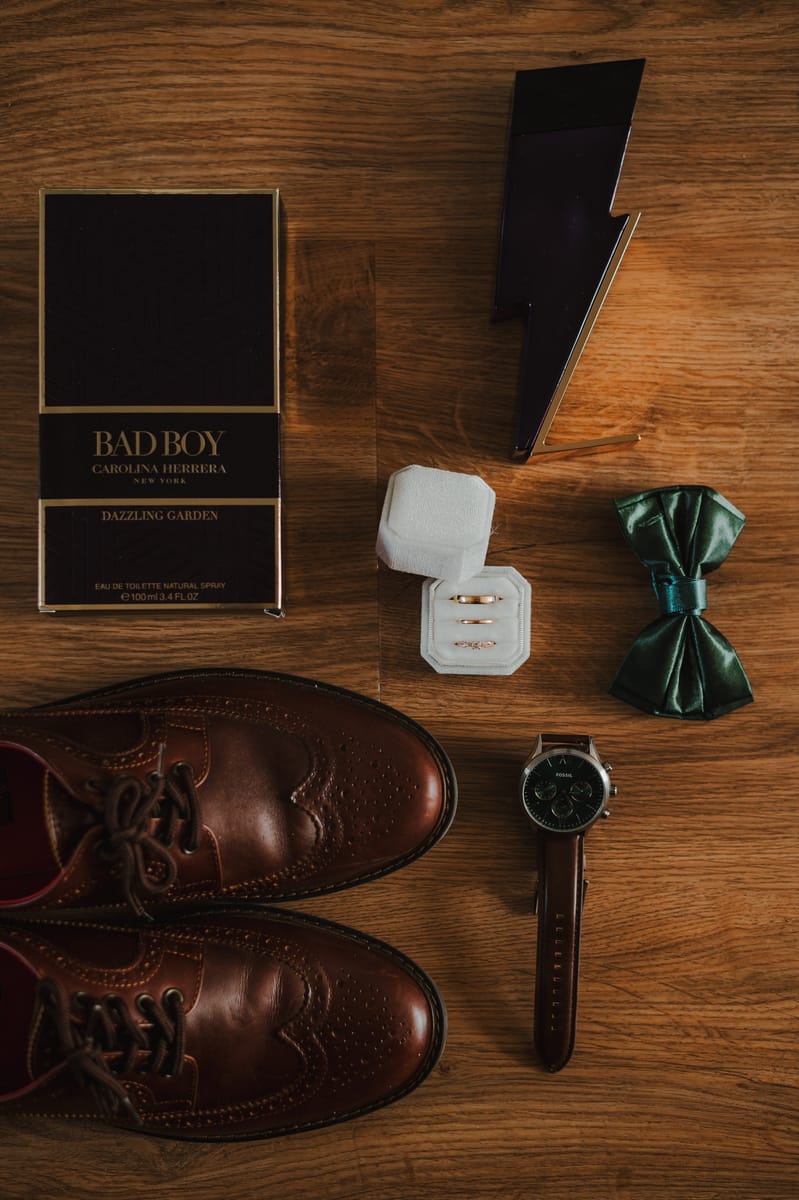 Detalles del novio vistos desde arriba: zapatos de piel, una cajita con anillos, un moño verde, un reloj y un perfume sobre madera