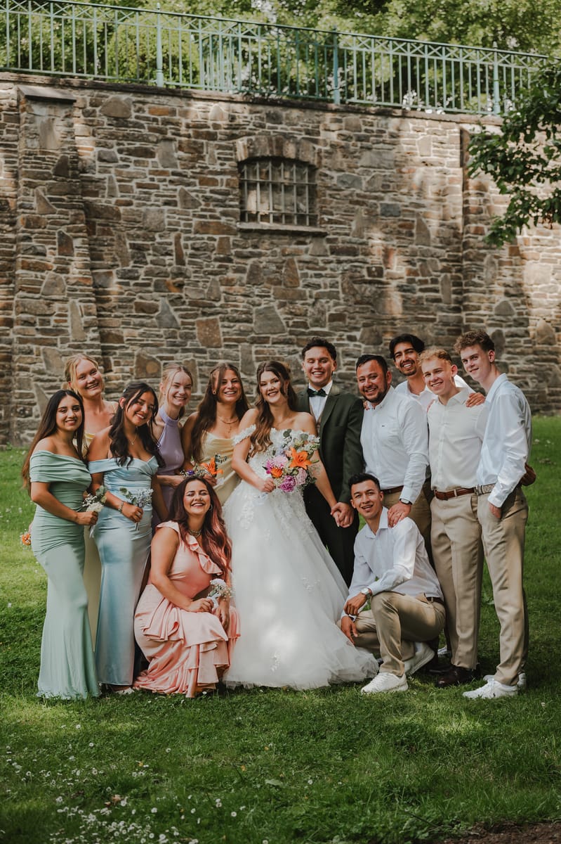 Retrato de grupo de los novios con sus amigas y amigos, sonriendo frente a un muro de piedra