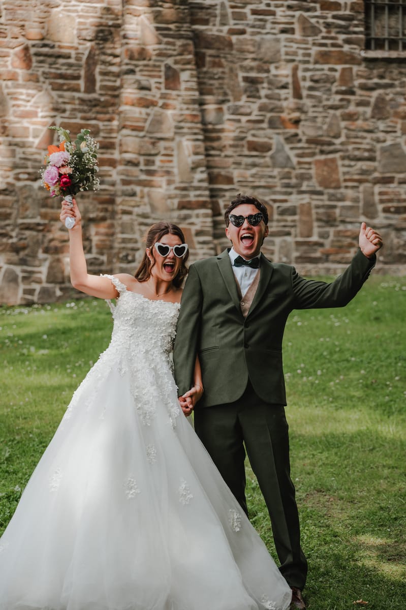 Los novios celebran con los brazos en alto y lentes de sol en forma de corazón, frente a un muro de piedra